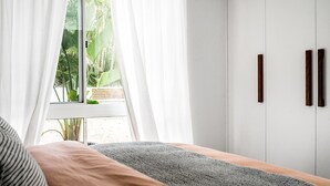 Une chambre à coucher avec un lit, une fenêtre avec des rideaux, une vue sur la verdure à l’extérieur et une armoire dotée de deux poignées sombres.