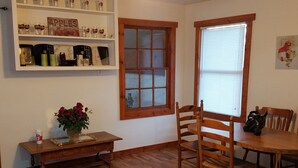 A room with wooden flooring, a dining table, a wooden chair, a small table with a vase of flowers, a shelf with various items, and a window with blinds.