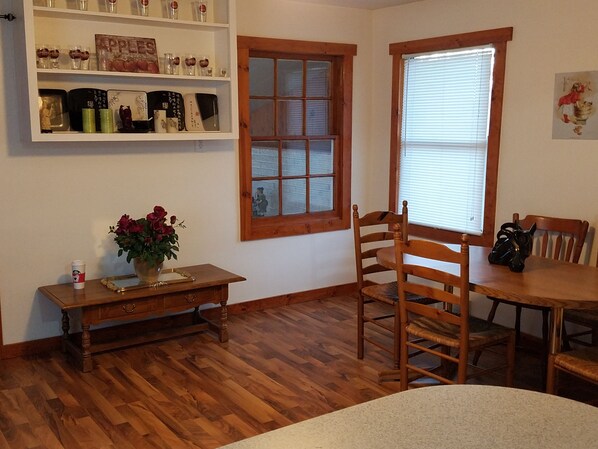 A room with wooden flooring, a dining table, a wooden chair, a small table with a vase of flowers, a shelf with various items, and a window with blinds.