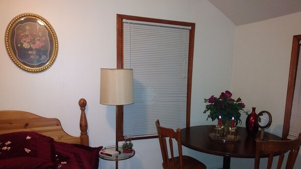 A bedroom with a bed, a bedside table with a lamp, a small table with chairs, a mirror, and a window with blinds.