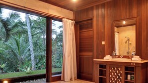 Un baño con tocador de madera, un espejo grande y vista a un jardín tropical a través de la puerta de cristal.