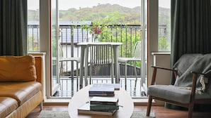 A living room with a sofa, a round coffee table with books, a chair, and a balcony with a view of greenery and hills. - Historic Townhouse in Clifden with courtyard+ balcony overlooking
Clifden Bay (Clifden)