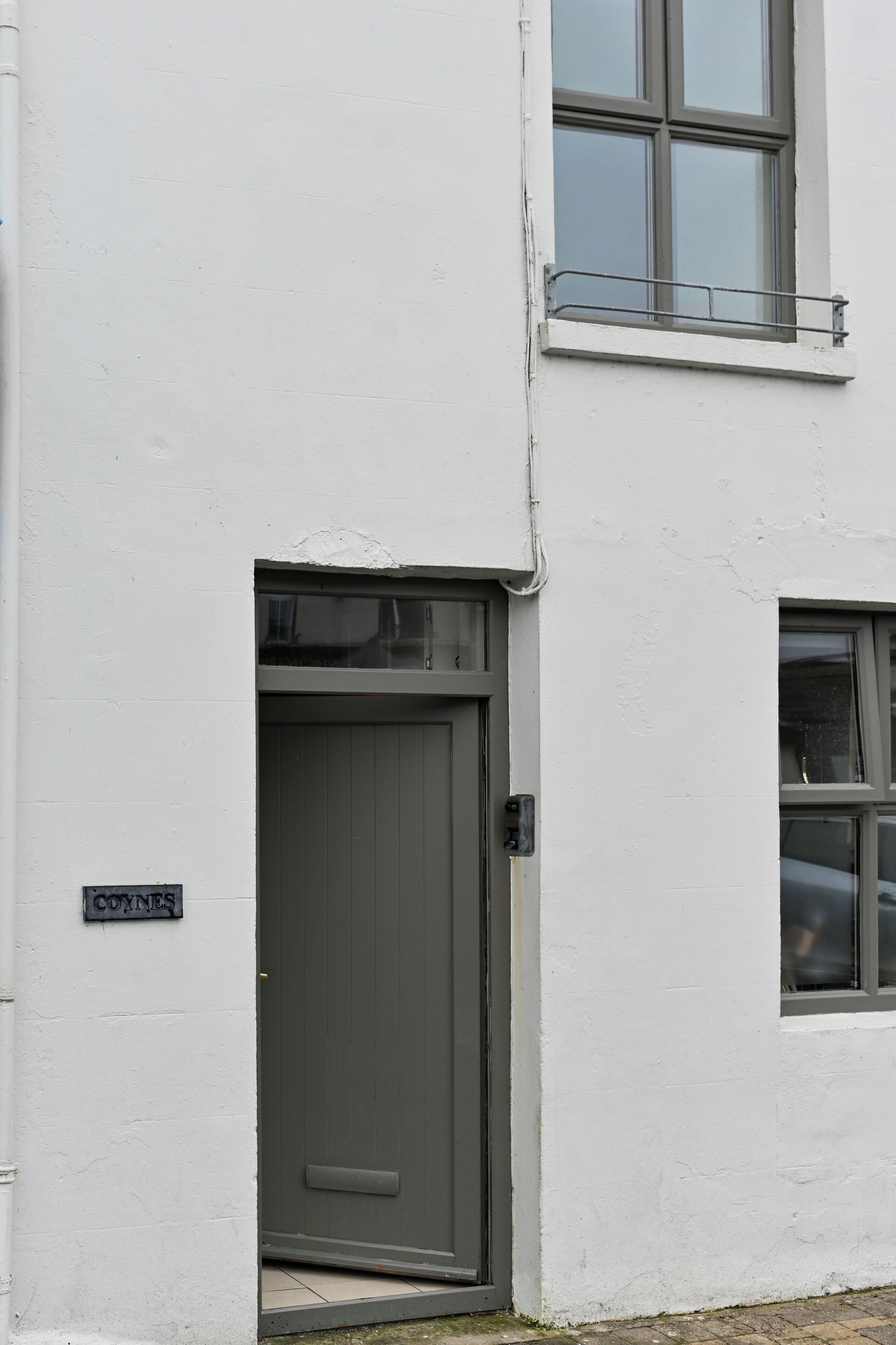 Un bâtiment blanc avec une porte grise et une fenêtre ornée d’une rambarde en métal.