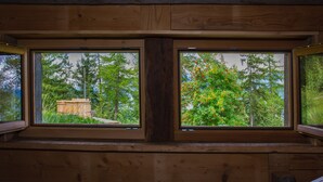 Eine Holzhütte mit zwei Fenstern, Holzwänden und Blick auf Bäume sowie eine Holzkonstruktion draußen.
