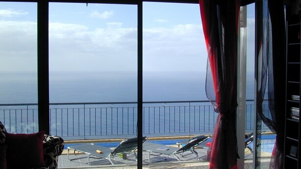 A room with a view of the sea through large windows, a balcony with sun loungers, and a patterned sofa.