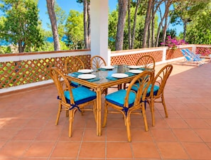 An outdoor dining area with a table set for four, surrounded by wicker chairs with blue cushions, on a terrace with a tiled floor and a lattice fence. -  Beachfront Villa with spacious garden and direct beach access  (San Sostene Marina)
