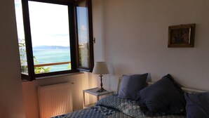 A bedroom with a bed, a nightstand with a lamp, a window with a view of the sea, and a painting on the wall. - "BLUE HOUSE" beautiful semi-detached villa with breathtaking views (PORTO SANTO STEFANO MONTE ARGENTARIO (GR)
