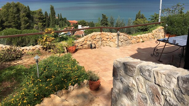 A terrace with a stone wall, potted plants, and a view of the sea and distant mountains. - "BLUE HOUSE" beautiful semi-detached villa with breathtaking views (PORTO SANTO STEFANO MONTE ARGENTARIO (GR)