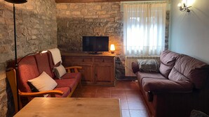 Una sala de estar acogedora con pared de piedra, vigas de madera en el techo, televisor y ventana con cortinas.