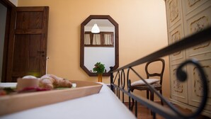 Une chambre avec une armoire en bois, un miroir, un lit recouvert d’une courtepointe blanche, une chaise et une petite table sur laquelle est posée une plante.