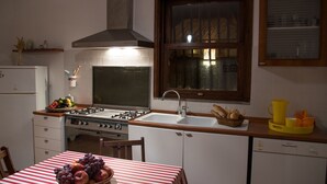 Une cuisine équipée d’une cuisinière à gaz, d’un évier et d’une fenêtre avec des rideaux. On y trouve une table avec un panier de fruits et une nappe à rayures.