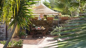 Une terrasse extérieure avec un mur en pierre, une table dressée avec des fruits et des boissons, et un parasol en chaume.