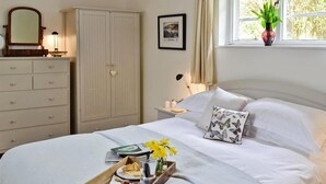 Ein aufgeräumtes Bett mit einem Frühstücksbrettchen, ein Nachttisch mit Lampe, ein Kleiderschrank und ein Fenster mit einer Pflanze.