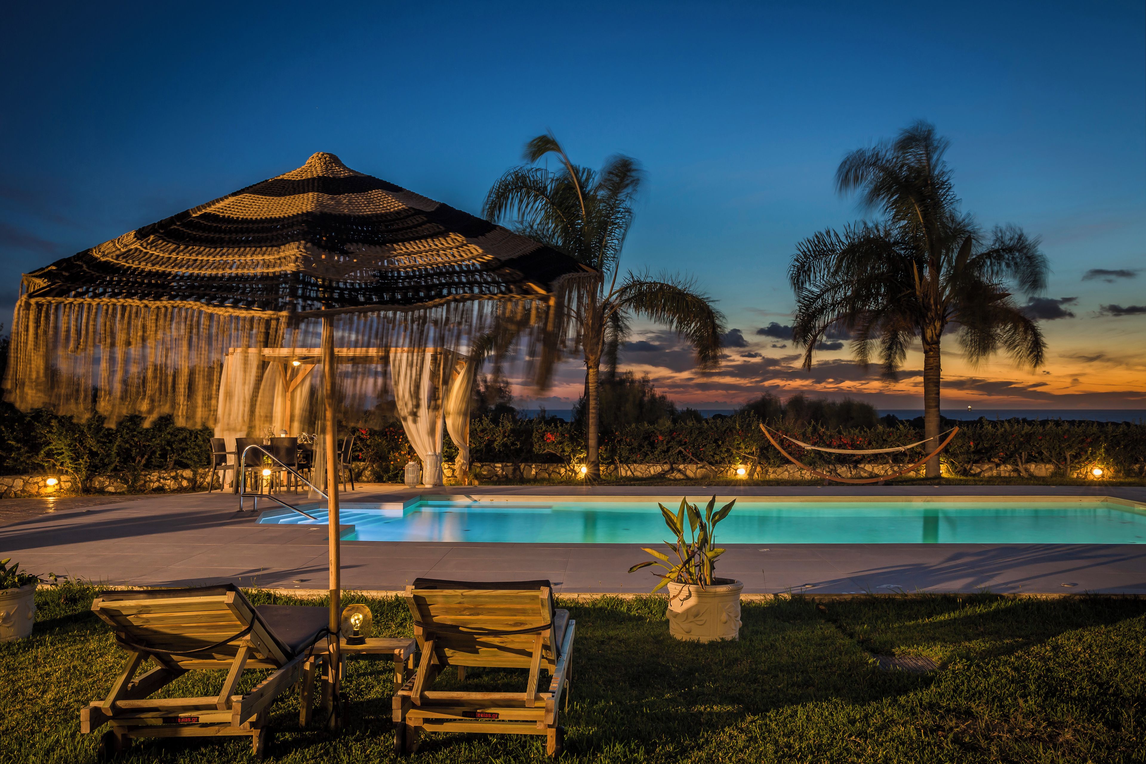 A pool area with lounge chairs, a gazebo, and palm trees at dusk.