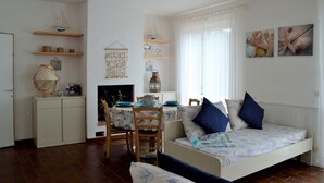 A cozy living room with a sofa, a dining table with chairs, and a fireplace. There are shelves with decorative items and a hanging net. - Apartment surrounded by tranquility with beautiful sea view (Capoliveri)