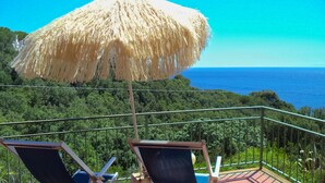 A balcony with two lounge chairs and a straw umbrella overlooking a forested area and the sea. - Apartment surrounded by tranquility with beautiful sea view (Capoliveri)