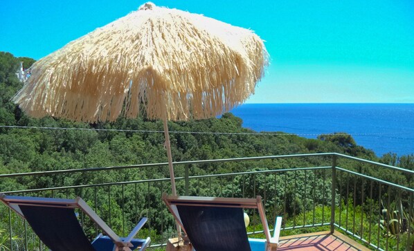 Varanda com duas cadeiras de descanso e um guarda-sol de palha, com vista para uma área arborizada e o mar.
