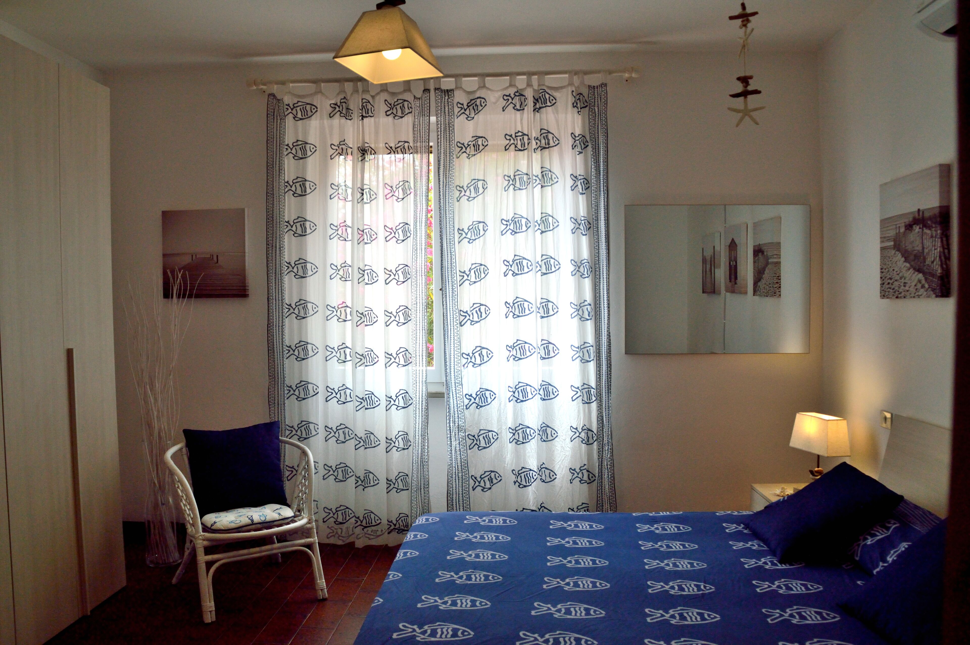A bedroom with a bed, a chair, a nightstand, a lamp, and curtains with a fish pattern.