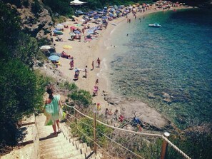 Ein Strand mit klarem Wasser, einer Treppe, die zum Ufer führt, und Menschen, die sich unter Sonnenschirmen entspannen.