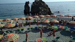 Una platja amb parasols i cadires a ratlles, formacions rocoses al fons, i persones nedant i relaxant-se.