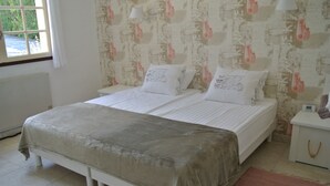 Cama de casal com colcha cinza, duas almofadas, mesa de cabeceira branca com abajur e papel de parede estampado.