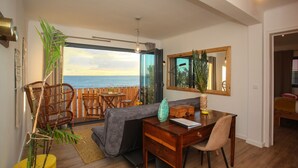Una habitación con piso de madera, un sofá gris, una mesa de comedor de madera con una silla, un espejo grande y vista al mar a través de una puerta de vidrio.