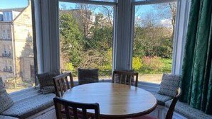 Un comedor con una mesa redonda y sillas, ventanas amplias con cortinas, y vistas a zonas verdes y edificios.