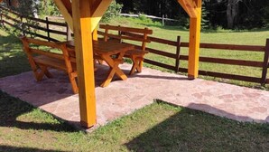 Um gazebo de madeira com telhado de metal, uma mesa para piquenique e um caminho de pedra.