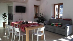 Ein Essbereich mit einem Holztisch, weißen Stühlen, einem grauen Sofa und einem an der Wand befestigten Fernseher.