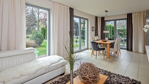Ett modernt vardagsrum med en vit soffa, en matplats med ett träbord och stolar samt stora fönster med utsikt över trädgården.