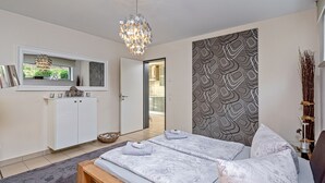 Ett modernt sovrum med en stor säng, en bänk, en kristallkrona, en garderob och en vägg med ett mönster.