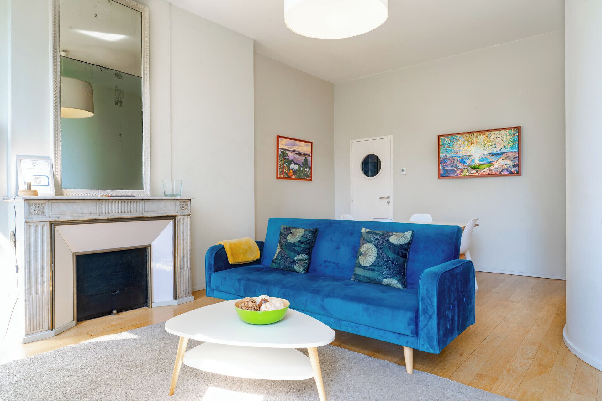 Una sala de estar con un sofá azul, una mesa de centro blanca, una chimenea y dos obras de arte enmarcadas en las paredes.
