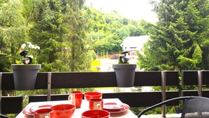 An outdoor seating area with a table set for dining, surrounded by potted plants and overlooking a lush green landscape with trees and a building in the distance.
