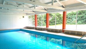 An indoor swimming pool with clear blue water, white tiled walls, and large windows offering a view of greenery outside.