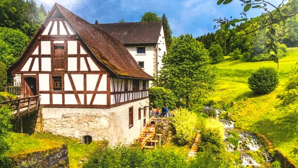 A traditional half-timbered house with a stone foundation, a wooden staircase leading to the entrance, and a small stream flowing nearby.