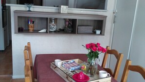 Ein Essbereich mit einem Holztisch, Stühlen und einer roten Tischdecke. Auf einem Tablett stehen ein Blumenstrauß und eine rosa Saucenflasche. Eine Ablage über dem Tisch beherbergt verschiedene Gegenstände.