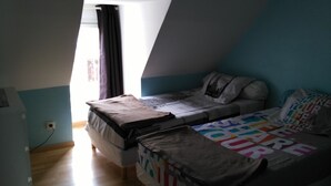 Ein kleiner, schummrig beleuchteter Raum mit schrägverlaufender Decke, einem Bett mit bunter Decke und einem Fenster mit Vorhang.