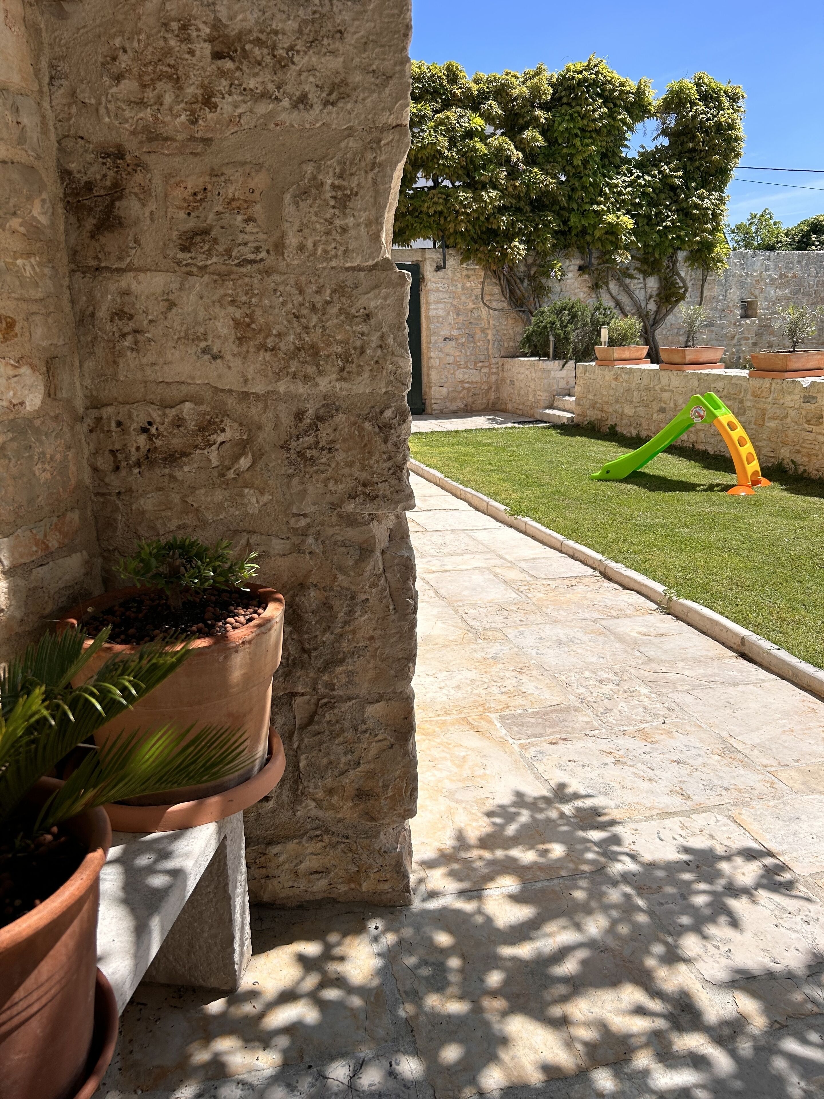 A stone wall with potted plants, a grassy area with a green and orange slide, and a clear blue sky.