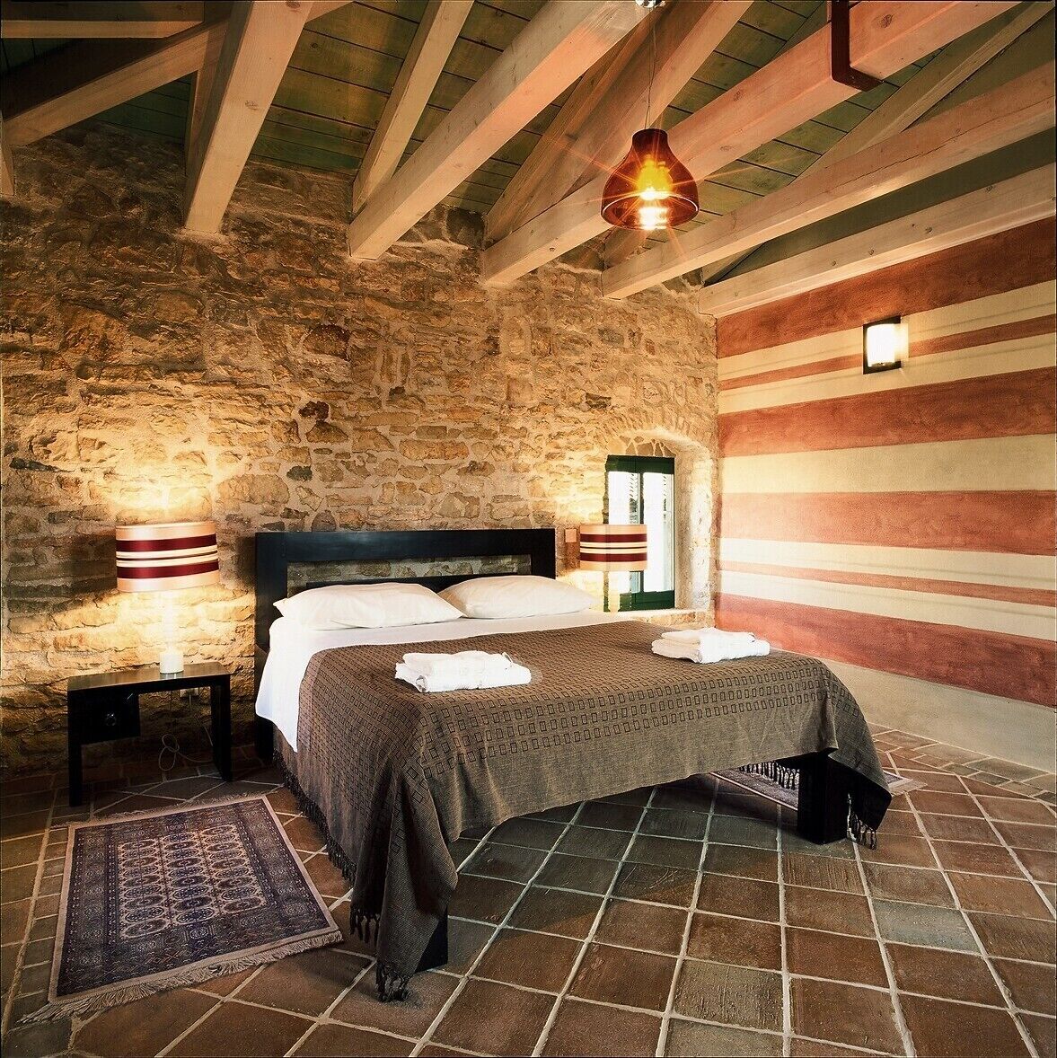 Una habitación con pared de piedra, una cama con colcha estampada, una mesita de noche con lámpara, y una lámpara colgante.