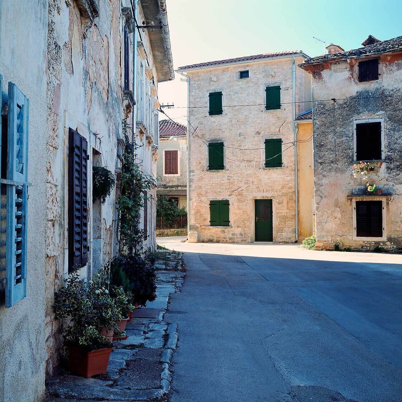 A narrow street with old stone buildings, some with shutters, and potted plants along the sidewalk.