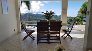 A room with a tiled floor, a wooden table with chairs, a vase with flowers, and a view of mountains and water.