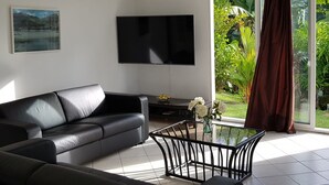 A modern living room with a black leather sofa, a flat-screen TV mounted on the wall, and a glass table with a metal frame.