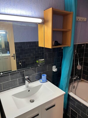 A bathroom with a white sink, a mirror, a wooden medicine cabinet, black tiled walls, and a shower area.