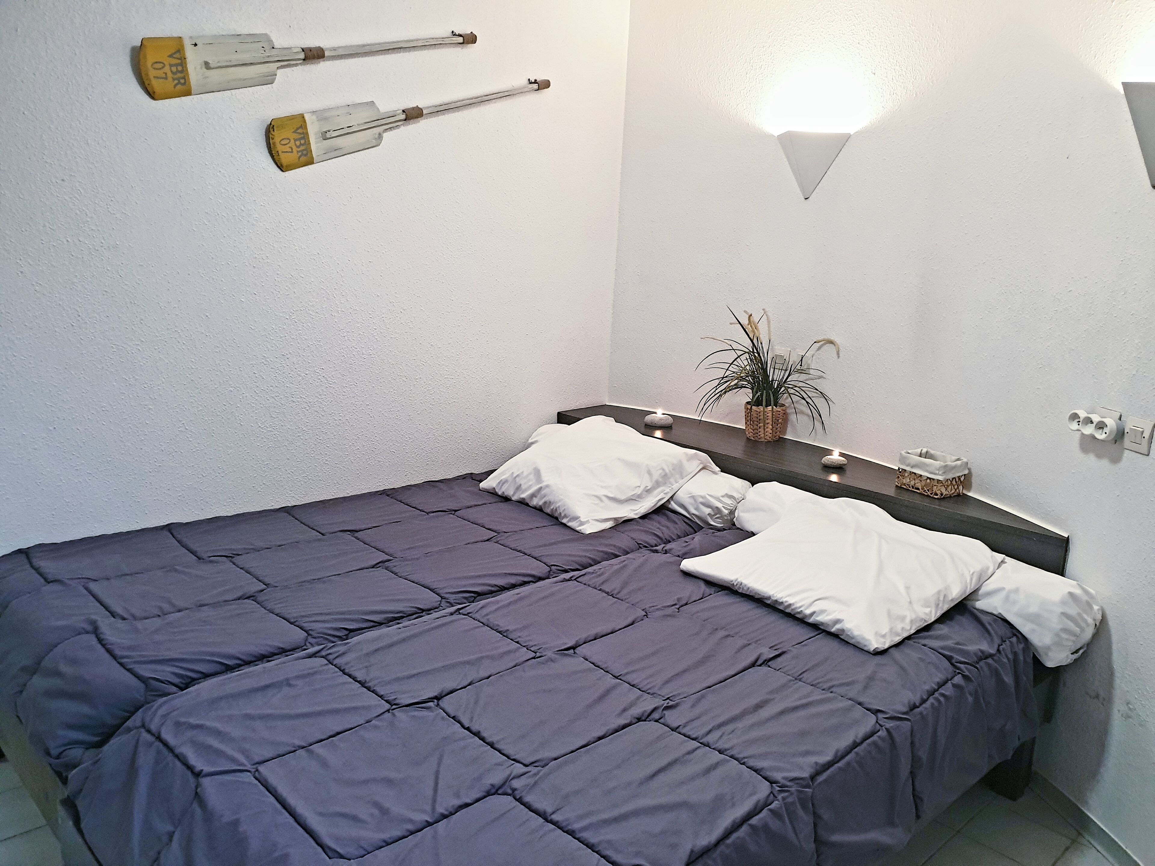 Una cama bien hecha con colcha azul oscuro y almohadas blancas, una mesita de noche con una planta y dos lámparas fijadas a la pared.