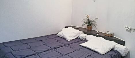 Una cama bien hecha con colcha azul oscuro y almohadas blancas, una mesita de noche con una planta y dos lámparas fijadas a la pared.