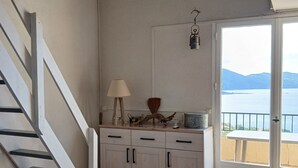 Une pièce avec un escalier, un canapé, un coin cuisine avec des placards, un luminaire suspendu et une vue sur la mer par une porte.