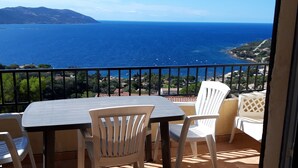 Un balcon avec une table et des chaises, offrant une vue sur la côte, la mer et les collines lointaines.