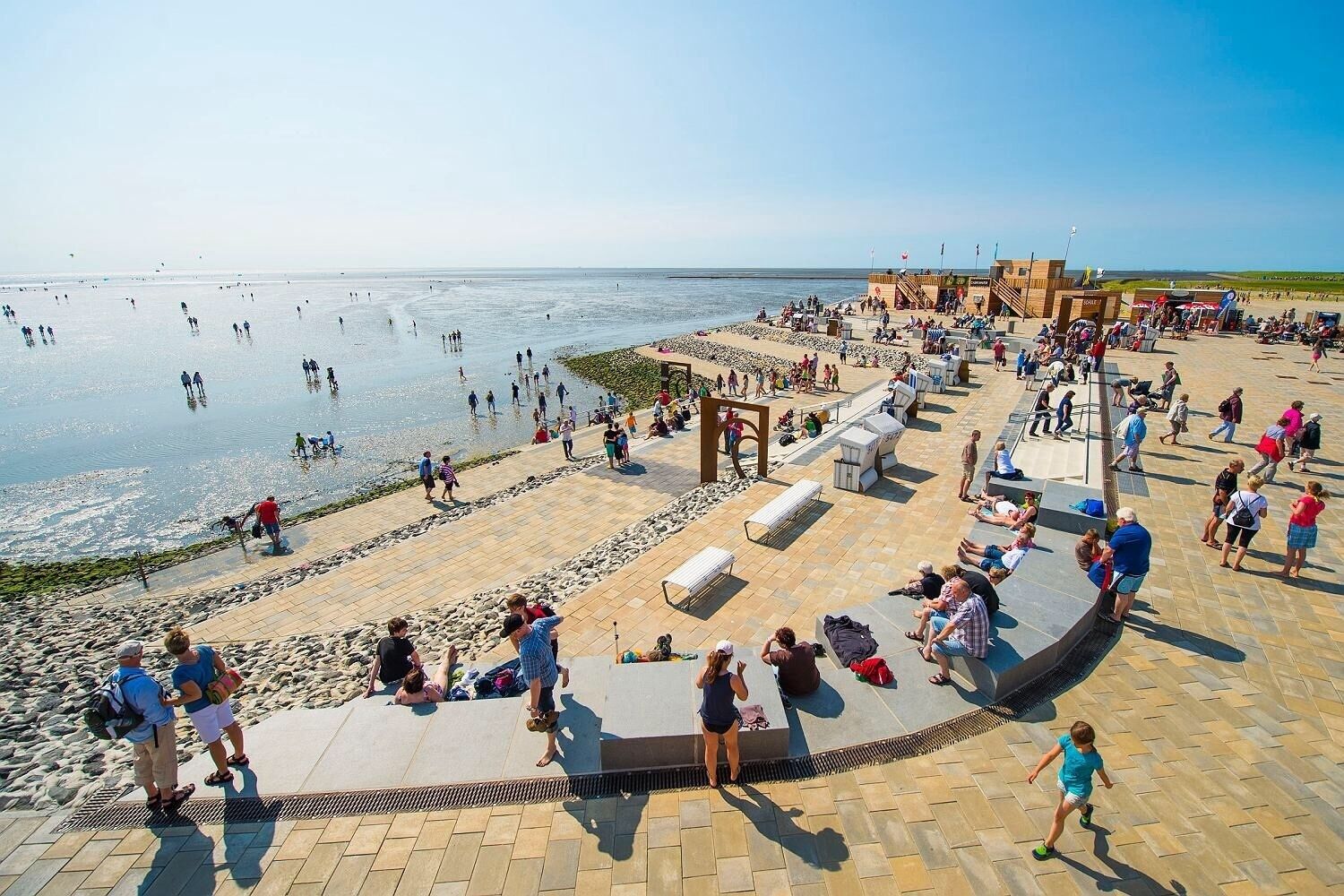 Une promenade en bord de mer, avec des escaliers en pierre menant à l’eau, des bancs et une structure en bois ornée de drapeaux.