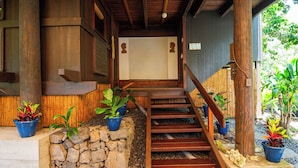 Un escalier en bois mène à une entrée abritée, agrémentée de plantes en pot et bordée d’un mur en pierre.