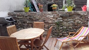 Un patio con un muro de piedra, una mesa redonda de madera, sillas de madera, una parrilla y plantas de colores en macetas.